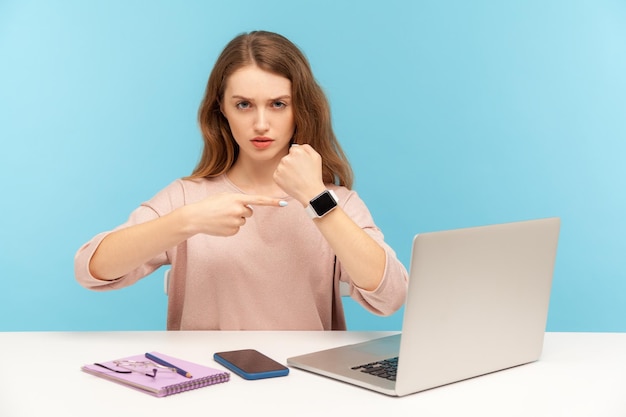 Dépêchez-vous de respecter la date limite Femme d'affaires irritée assise sur le lieu de travail et pointant la montre-bracelet regardant avec colère l'avertissement de l'heure tardive pour travailler en studio intérieur isolé sur fond bleu