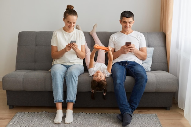 Dépendance aux gadgets Jeune famille de trois personnes tenant et utilisant différents appareils électroniques assis sur un canapé Parents et leur fille avec des gadgets modernes exprimant des émotions positives