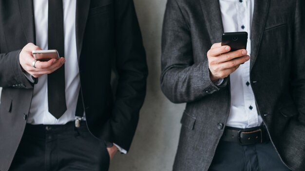 Dépendance aux réseaux sociaux. Temps de pause de la vie d'entreprise moderne. Deux hommes en costume d'affaires s'appuyant sur le mur, utilisant des smartphones.