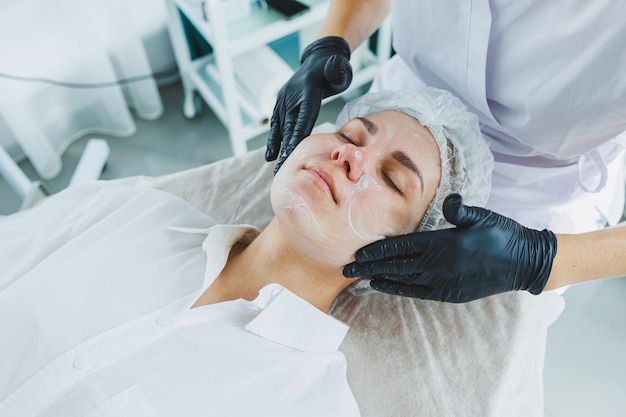 Un dermatologue traite le visage d'une femme allongée sur une table de massage Soin de la peau du visage Une esthéticienne applique un masque au visage de la femme