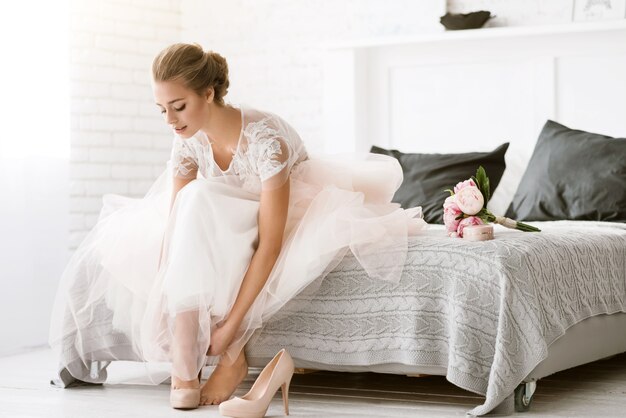 Les dernières minutes. Charmante jeune mariée ravie assise sur le lit dans une chambre de couleur blanche tout en portant ses chaussures et exprimant sa tendresse