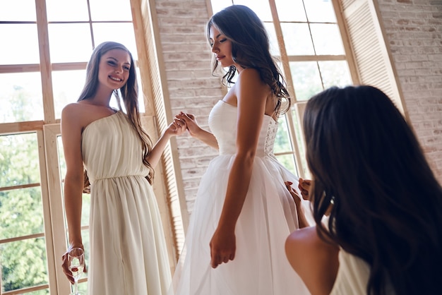 Derniers détails. Deux jeunes femmes attirantes aidant la belle mariée à s'habiller
