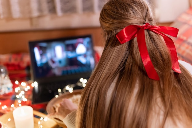 Derrière la tête d'une femme avec de longs cheveux blonds et une vue arrière de l'arc rouge en regardant le film de Noël sur un ordinateur portable à la maison de camping dans un lit confortable avec une guirlande de lumières rougeoyantes la nuit à Noël ou au réveillon du nouvel an