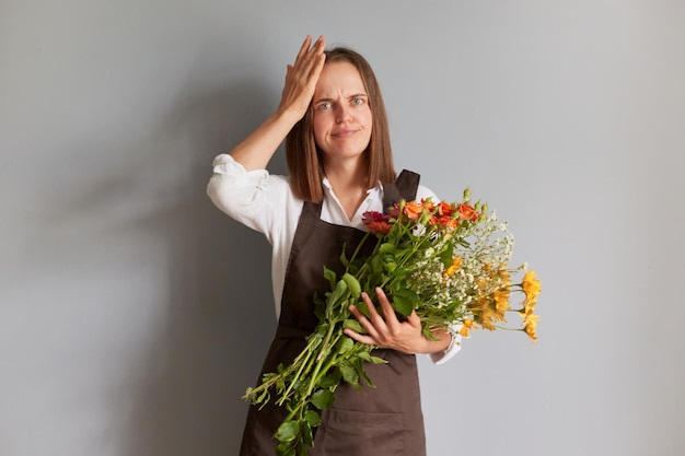 Désespoir triste bouleversée femme fleuriste portant un tablier marron tenant des fleurs isolées sur fond gris montrant un geste facepalm ayant des problèmes avec son entreprise