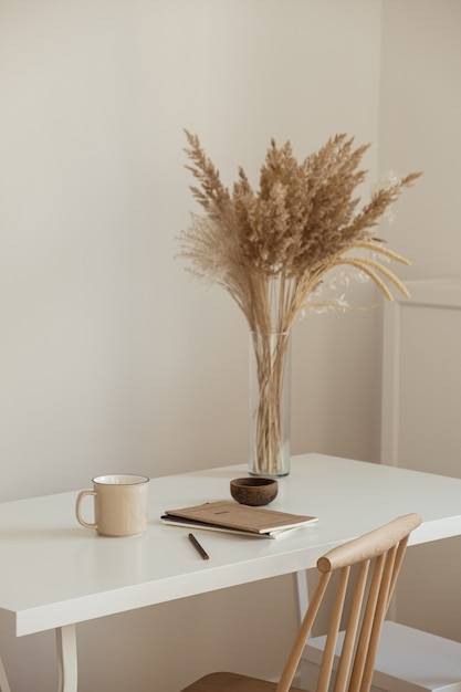 Design d'intérieur d'espace de travail de bureau minimal esthétique. Tasse, cahier, bouquet floral d'herbe de pampa sur la table blanche contre le mur blanc