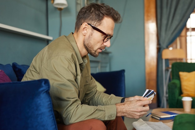 Designer mature en lunettes assis sur un canapé et examinant les motifs colorés dans ses mains au bureau