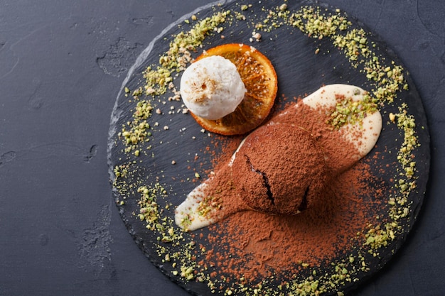 Dessert français classique. Gâteau de lave fondant au chocolat chaud avec crème pâtissière et glace à la vanille, pépites de cacao sur ardoise, tranche d'orange et crumble de pistache, fond noir, vue de dessus, espace de copie