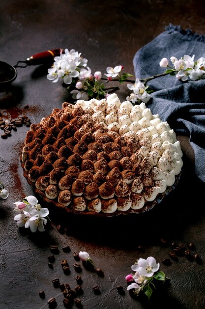 Dessert italien traditionnel tiramisu sans gluten fait maison saupoudré de poudre de cacao décoré de pommier en fleurs, de serviette textile bleue et de grains de café sur une surface de texture sombre.