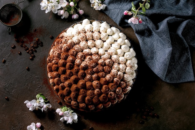 Dessert italien traditionnel tiramisu sans gluten fait maison saupoudré de poudre de cacao avec pommier en fleurs, serviette textile bleue et grains de café sur une surface de texture sombre. Vue de dessus, mise à plat