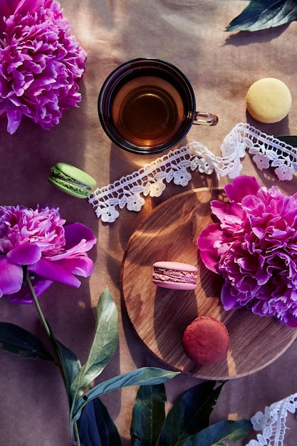 Dessert de macarons lumineux et tisane parmi des fleurs de pivoine rose en fleurs sous des ombres dures à la mode sur la terrasse Relaxation esthétique estivale méditative réfléchie mode de vie de bonne humeur