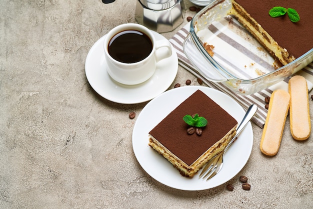 Dessert tiramisu italien traditionnel dans un plat de cuisson en verre et portion sur table en béton gris