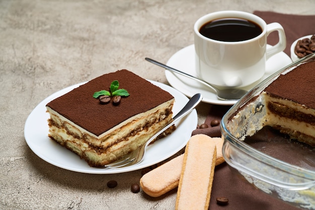 Dessert tiramisu italien traditionnel dans un plat de cuisson en verre et portion sur table en béton gris