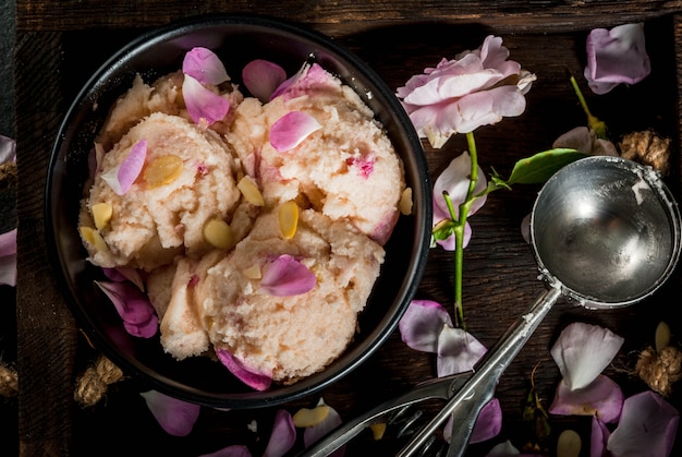 Desserts rafraîchissants d'été Alimentation végétalienne Crème glacée avec des pétales de rose et des tranches d'amandes sur un vieux plateau en bois sur une table en pierre noire Avec une cuillère pour la crème glacée et les ingrédients