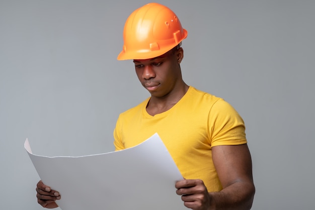 Dessin de plan directeur. Sérieux jeune homme à la peau sombre attentif en casque considérant le dessin de plan tenant dans les mains