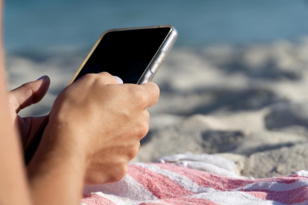 Détail des mains d'une femme aux ongles peints tenant son smartphone sur la plage