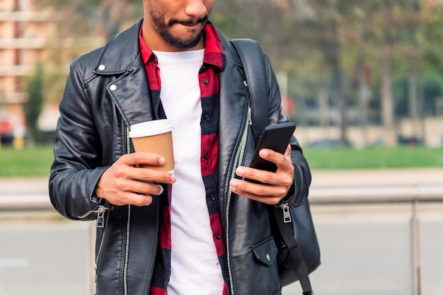 Détail des mains d'un jeune étudiant latin élégant consultant son téléphone avec un café à emporter, concept de technologie et mode de vie urbain