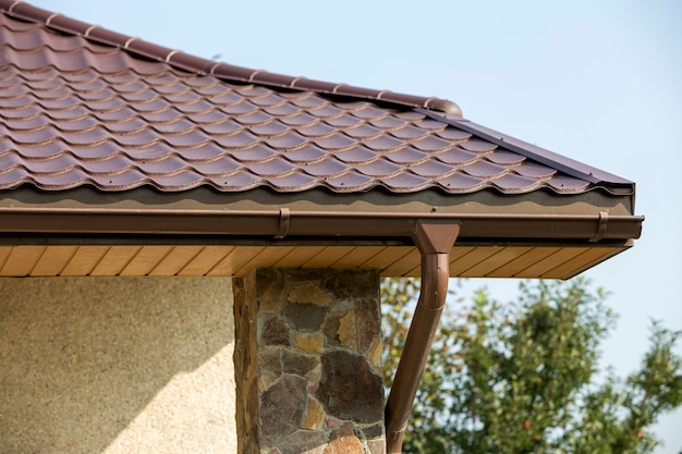 Détail d'un nouveau coin de maison moderne avec des murs en stuc décoré de pierres naturelles, un toit en bardeaux marron et un système de tuyaux de gouttière sur le ciel bleu. Immobilier .