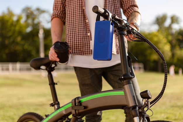 Photo détails de l'homme élégant hipster dans le parc écoutant de la musique sur un haut-parleur sans fil à l'aide d'un vélo électrique