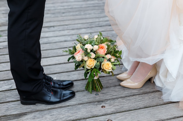 Détails de mariage: chaussures classiques noires et beiges de la mariée et du marié. Bouquet de roses se tenant sur le sol entre elles. Les nouveaux mariés debout devant l'autre