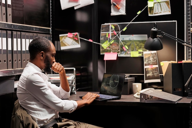 Détective privé regardant des photos de caméra de vidéosurveillance sur un ordinateur portable tout en étant assis au bureau. Enquêteur afro-américain analysant des photographies de surveillance sur écran d'ordinateur