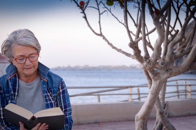 Détendez-vous et prenez votre retraite pour une femme âgée profitant du coucher de soleil en lisant un livre assis près de la plage et en mangeant une pomme. Horizon au-dessus de l'eau.