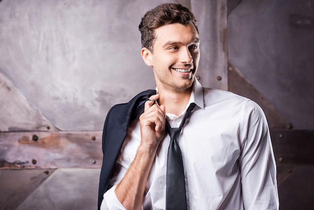 Détendu beau. Beau jeune homme en chemise et cravate portant sa veste sur l'épaule et regardant loin avec le sourire en se tenant debout sur fond de métal