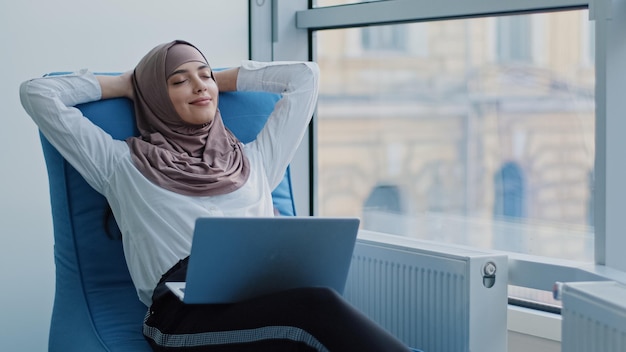 Détendue belle travailleuse arabe musulmane assise dans un fauteuil confortable méditant une femme arabe calme et positive se détendre tenir la main derrière la tête rêver se reposer de l'ordinateur s'étirer ressentir la tranquillité d'esprit