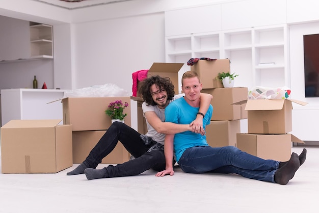 Détente dans une nouvelle maison. Enthousiaste jeune couple gay assis sur le sol tandis que des boîtes en carton s'étendent tout autour d'eux