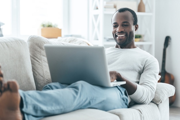 Détente à la maison. Beau jeune homme africain regardant un ordinateur portable et souriant en position couchée sur le canapé à la maison