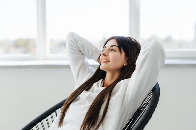 Détente totale Jolie jeune femme tenant les mains derrière la tête et souriant alors qu'il était assis à la chaise à la maison