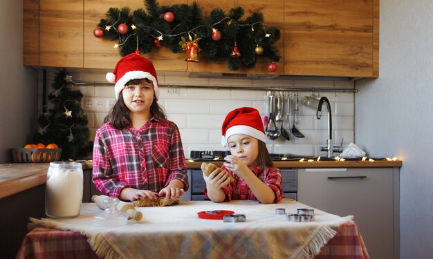 Deux adolescentes en chemises à carreaux rouges et chapeaux de Noël font des biscuits au gingembre avec de la pâte et rient joyeusement.
