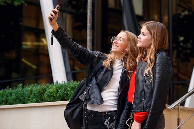 Deux adolescentes élégantes et heureuses avec smartphone faisant selfie près d'un bâtiment moderne et élégant dans la rue.