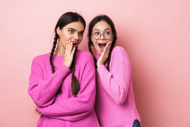 deux adolescentes heureuses avec des tresses exprimant l'émerveillement tout en regardant la surface isolée sur le mur rose