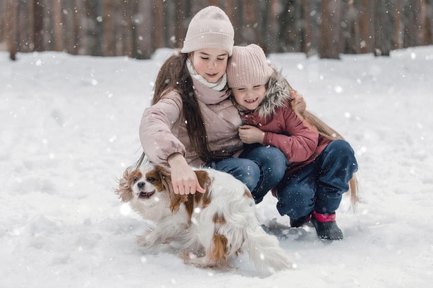 Deux adorables jeunes filles s'amusant ensemble par la belle forêt du parc gelé Sœurs mignonnes jouant dans la neige avec un chien