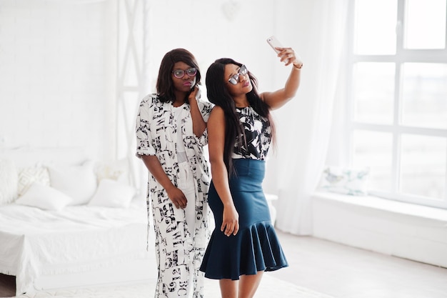 Deux amies africaines portent des lunettes posées dans une salle blanche intérieure et font du selfie par téléphone portable