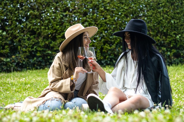 Deux amies en chapeaux assis sur l'herbe en train de griller avec des verres de vin.