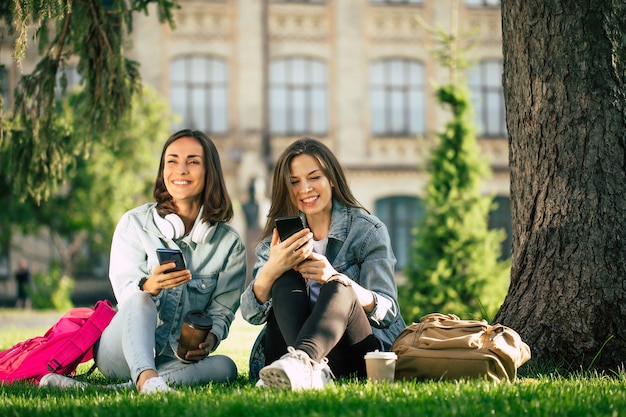 Deux amies étudiantes heureuses et excitées en vêtements décontractés se détendent dans le parc universitaire avec des smartphones et s'amusent.