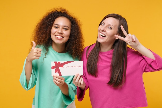 Deux amies européennes filles américaines en vêtements verts roses posant isolés sur fond jaune. Concept de style de vie des gens. Maquette de l'espace de copie. Tenez un certificat-cadeau montrant le signe de victoire du pouce vers le haut.