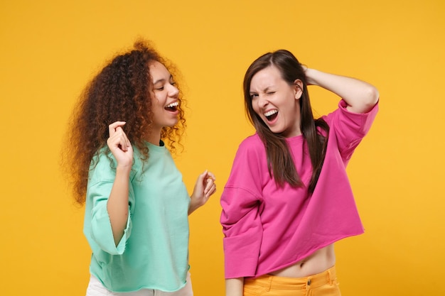 Deux amies gaies riant filles européennes et afro-américaines en vêtements verts roses posant isolées sur le portrait de studio de fond orange jaune. Concept de style de vie des gens. Maquette de l'espace de copie.