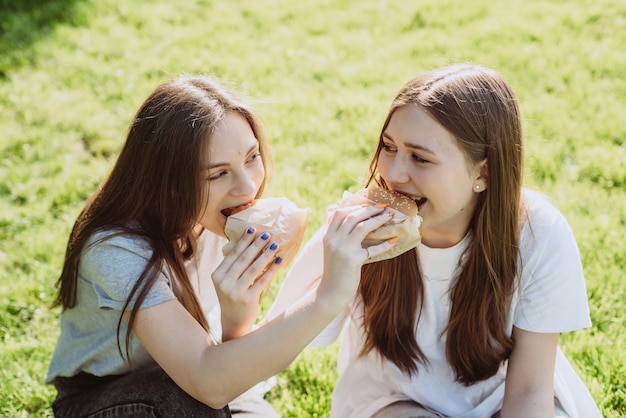 Deux amies de jeunes femmes mangeant de délicieux hamburgers dans le parc sur l'herbe. Pas une alimentation saine. Mise au point sélective douce