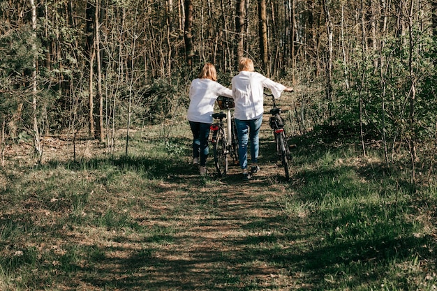 Deux amies matures marchent le long de la route avec des vélos en forêt. Vue arrière