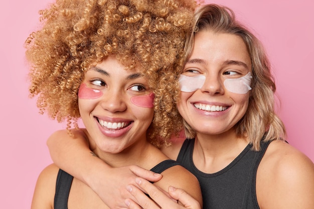 Deux amies positives s'embrassent avec amour appliquent des patchs d'hydrogel sous les yeux sourient largement vêtus de t-shirts noirs isolés sur fond rose Bien-être beauté et soins de la peau