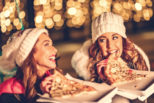 Deux amies profitent de la soirée, se regardent et rient avec des tranches de pizza dans leurs mains.