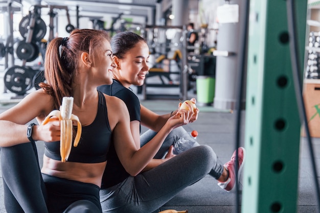 Deux amies vêtues de vêtements de sport sont dans la salle de gym en train de manger des fruits et de faire une pause.