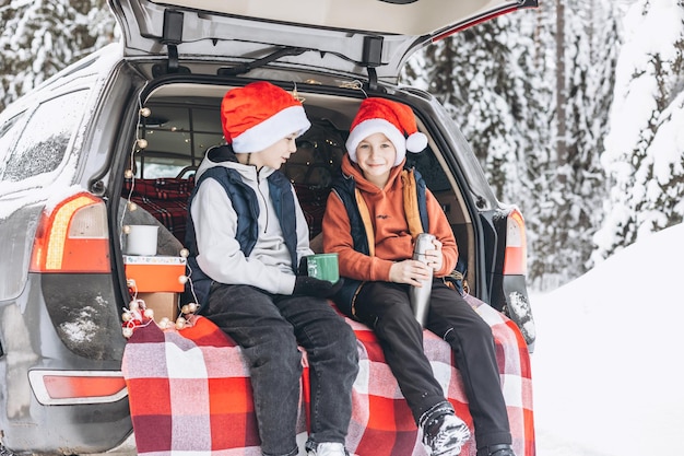 Deux amis adolescents garçons en bonnets rouges avec thermos assis dans le coffre d'une voiture décorée pour Noël Aventure sur la route et concept de voyage local Pique-nique d'hiver dans la forêt enneigée Vacances du nouvel an