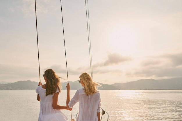 Deux amis en croisière de vacances ensemble tenant des câbles de gréement profitant du coucher de soleil Deux femmes profitant du coucher de soleil ensemble tenant des câbles de gréement de bateau