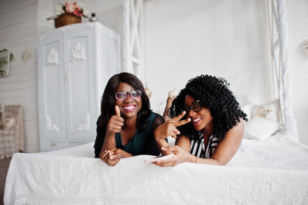 Deux amis de femme africaine portent des lunettes allongé sur le lit à l'intérieur de la salle blanche et regardant le téléphone mobile. Ils montrent le pouce.