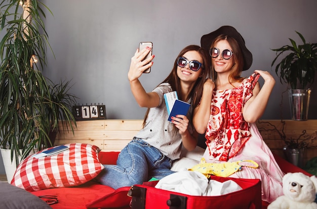 Deux amis gais heureux filles portent des lunettes de soleil faisant selfie assis sur le lit avec des vêtements et valise concept de voyage à l'intérieur