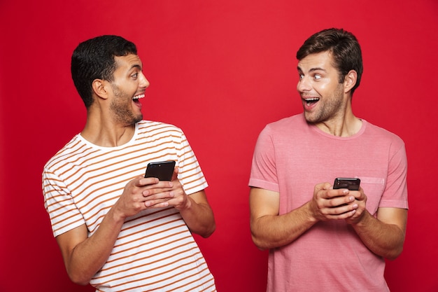 Deux amis hommes gais debout isolé sur mur rouge, à l'aide de téléphones mobiles