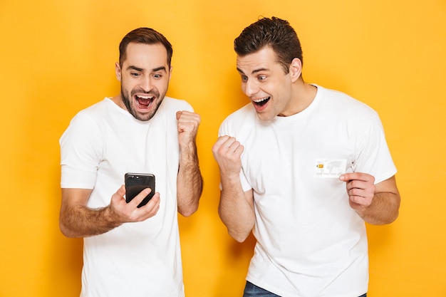 Deux amis joyeux et excités portant des t-shirts vierges isolés sur un mur jaune, regardant un téléphone portable, célébrant le succès